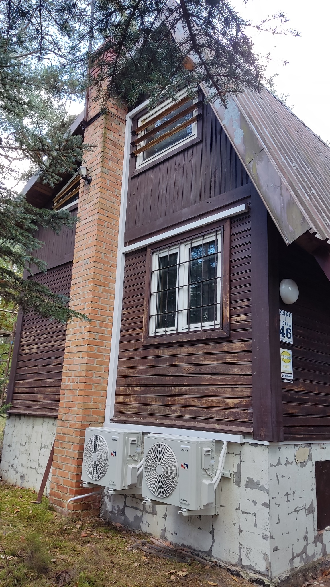 Dwa białe agregaty klimatyzacyjne zamontowane na ścianie drewnianego domu z ceglanym kominem i oknami z kratami.