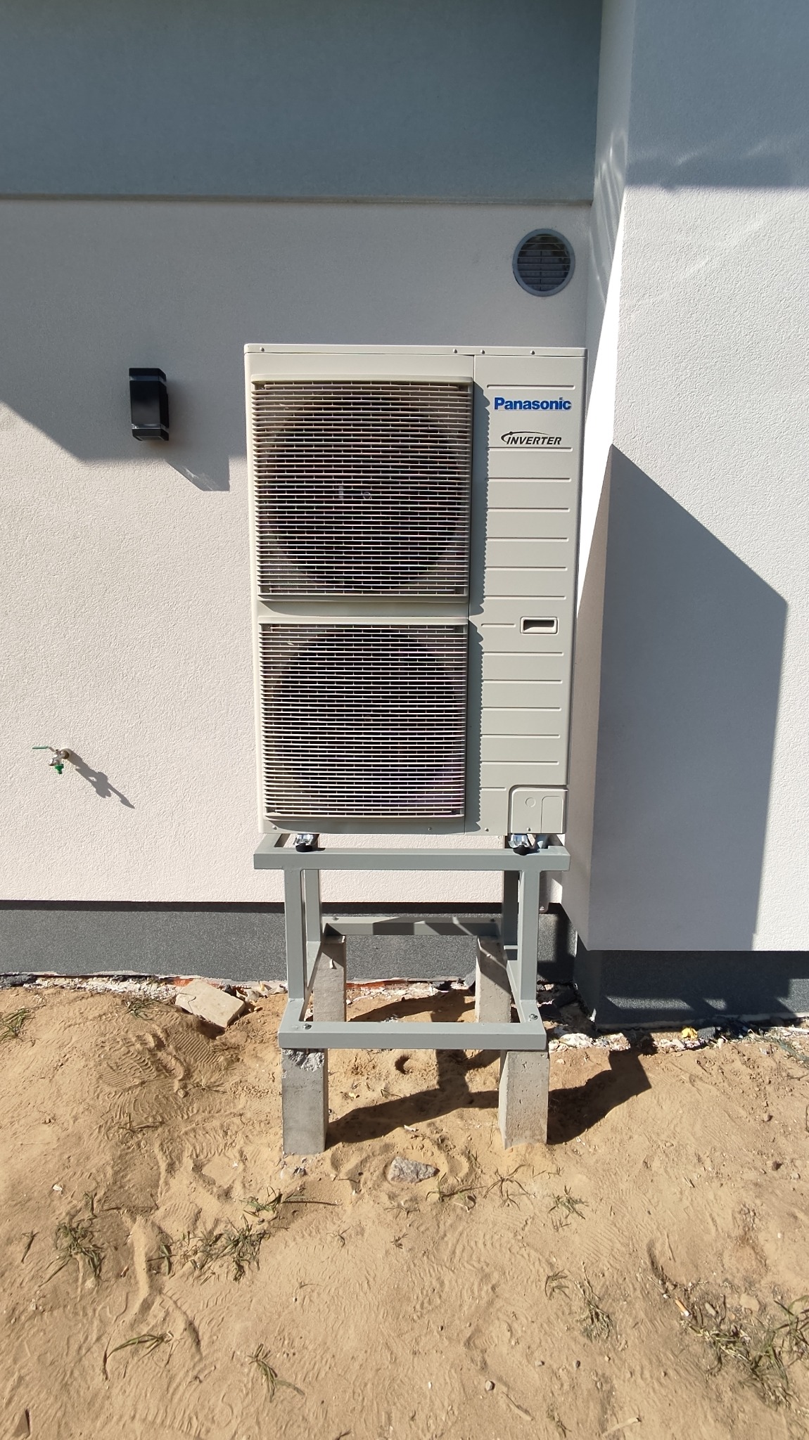 Zewnętrzna jednostka pompy ciepła Panasonic Inverter zamontowana na metalowym stelażu z betonowymi podstawami, przy białej elewacji budynku z widocznym punktem poboru wody i okrągłym otworem...