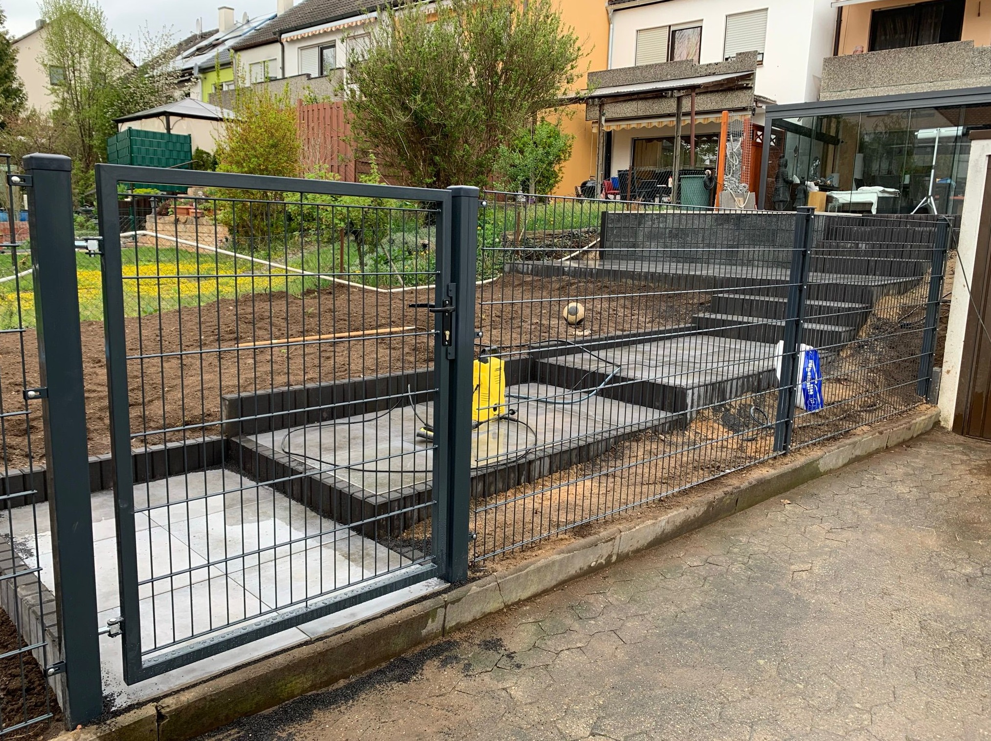 Metalowa brama ogrodzeniowa w kolorze grafitowym, prowadząca do ogrodu z przygotowanymi fundamentami pod taras z kostki, widoczny sprzęt budowlany.