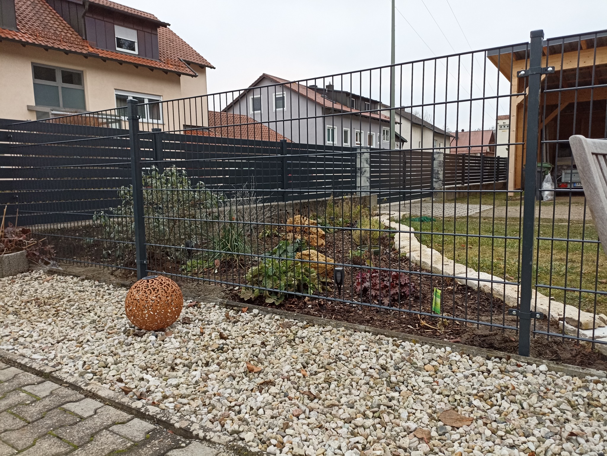 Nowoczesne ogrodzenie panelowe w kolorze antracytowym oddziela ogród z kamienną ścieżką i ozdobną kulą od posesji z zabudową jednorodzinną w tle.