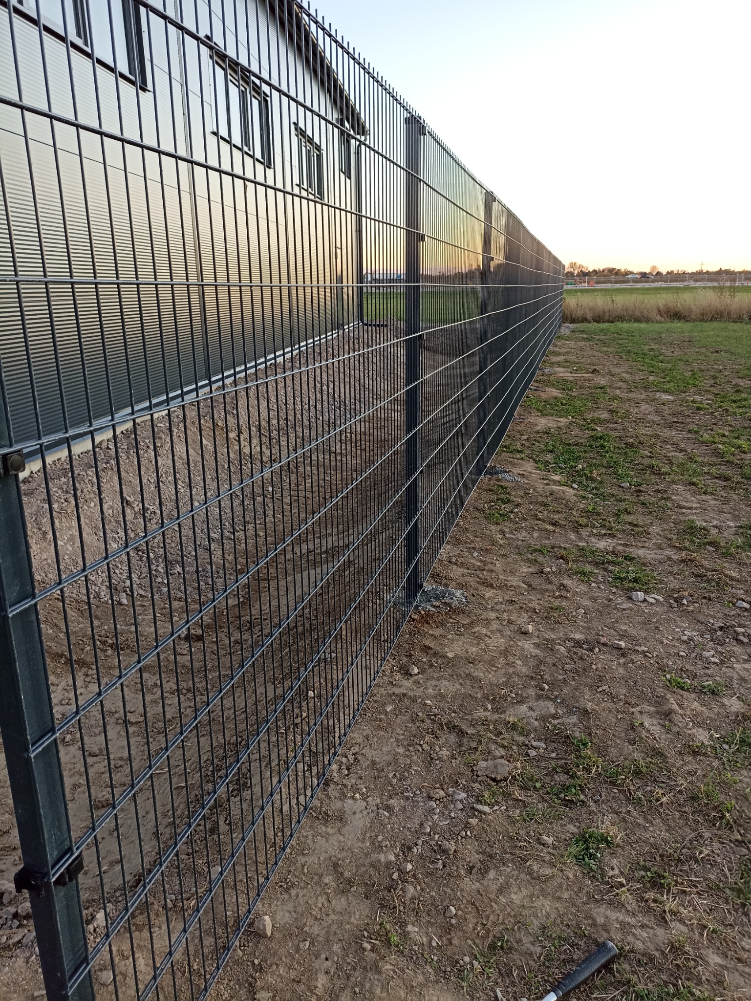 Metalowa siatka ogrodzeniowa, widok z boku, z budynkiem przemysłowym w tle, świeżo usypana ziemia przy podstawie ogrodzenia.