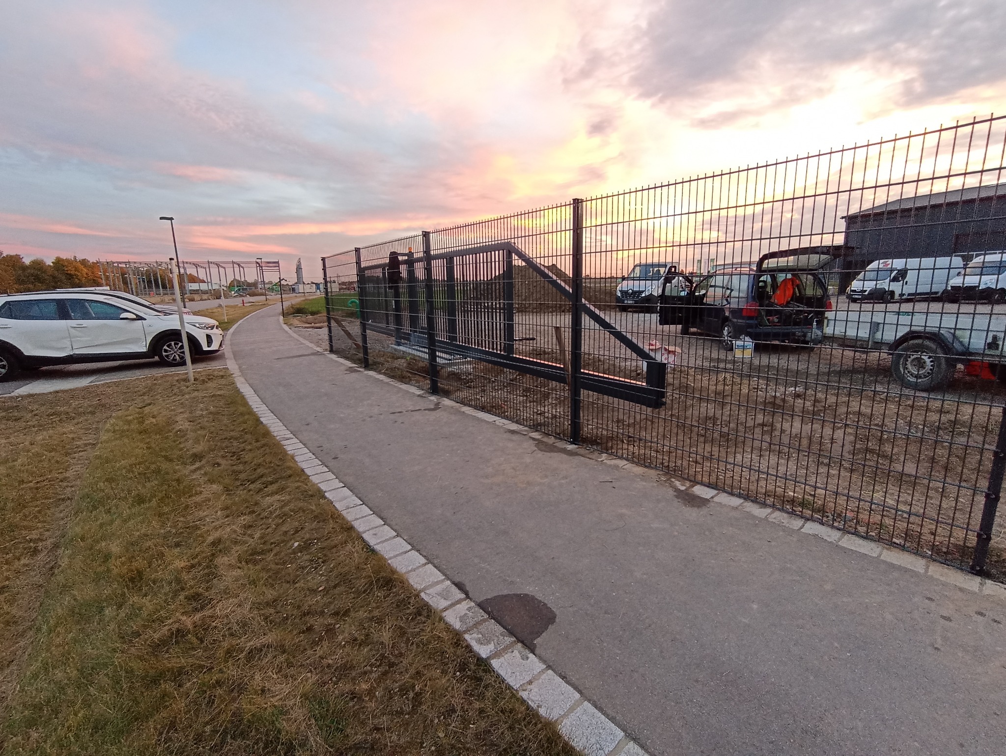 Nowo zainstalowana przesuwna brama wjazdowa z siatki stalowej, widoczna z boku ścieżki, z samochodami dostawczymi w tle i malowniczym zachodem słońca.