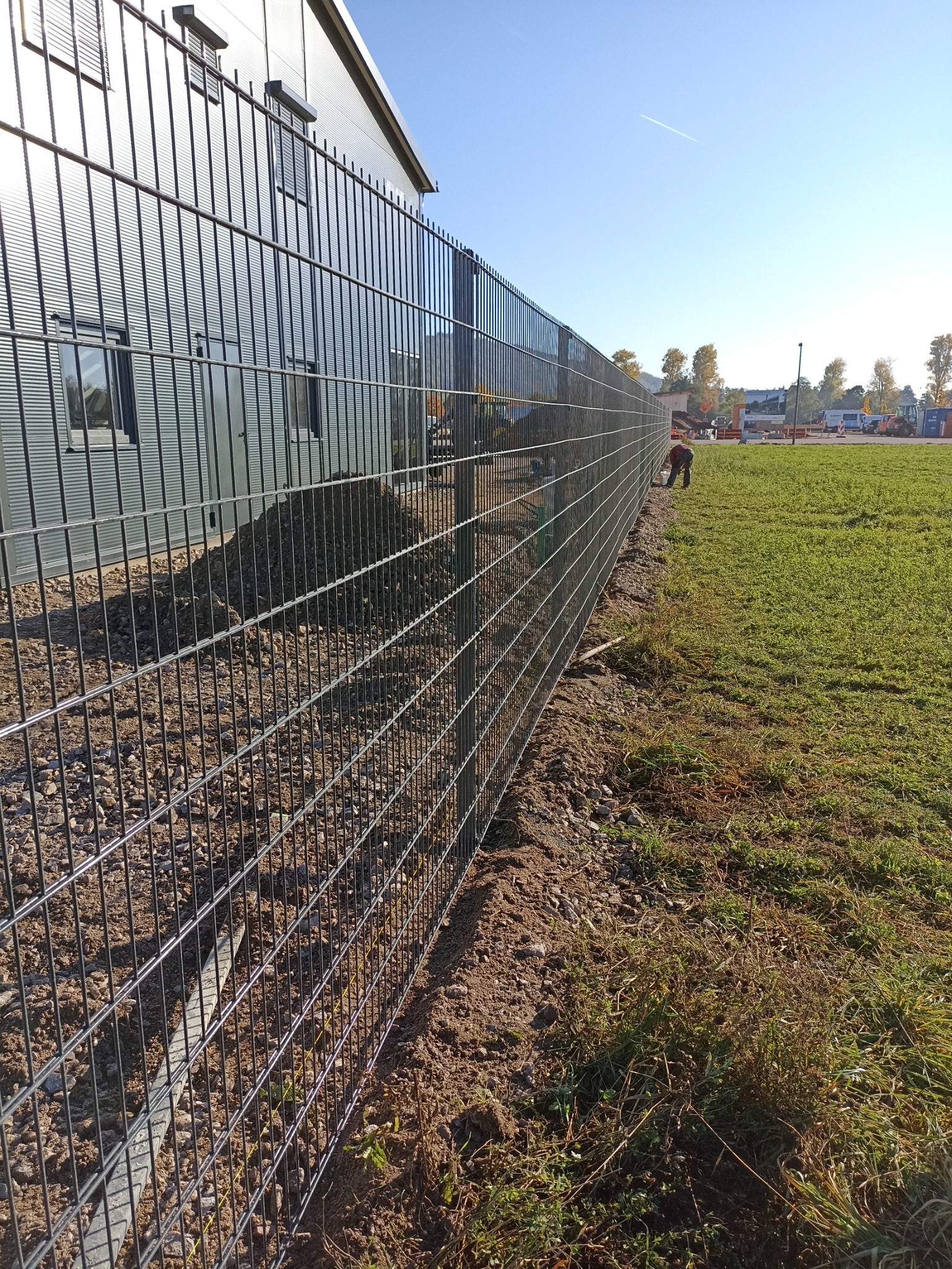 Montaż ogrodzenia panelowego zgrzewanego przy budynku przemysłowym, widoczna świeżo usypana ziemia i pracownik przy stabilizacji słupka.