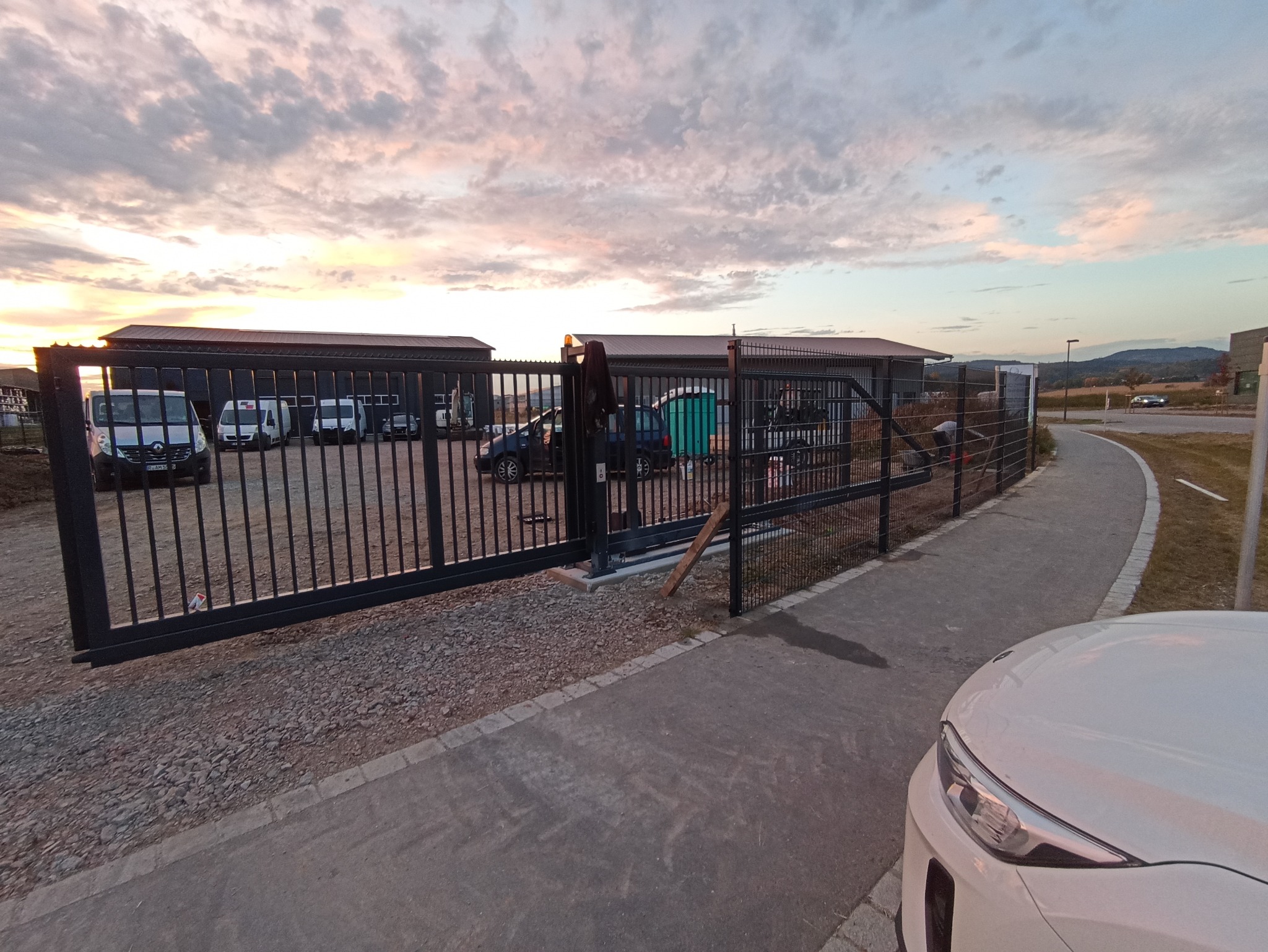 Czarna, metalowa brama przesuwna z siatką w tle, częściowo otwarta, z widocznym mechanizmem, na tle budynków gospodarczych, samochodów dostawczych i zachodzącego słońca.