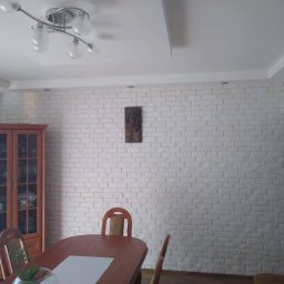 Shp-Rem - Biała ściana z cegieł dekoracyjnych w jadalni, oświetlona lampą sufitową, z drewnianymi meblami i dekoracją ścienną.