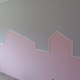 Shp-Rem - Ściana w pokoju dziecięcym pomalowana na szaro i różowo, z dekoracyjnym obramowaniem w kształcie domków, tworzącym iluzję panoramy miasta.