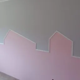 Ściana w pokoju dziecięcym pomalowana na szaro i różowo, z dekoracyjnym obramowaniem w kształcie domków, tworzącym iluzję panoramy miasta.