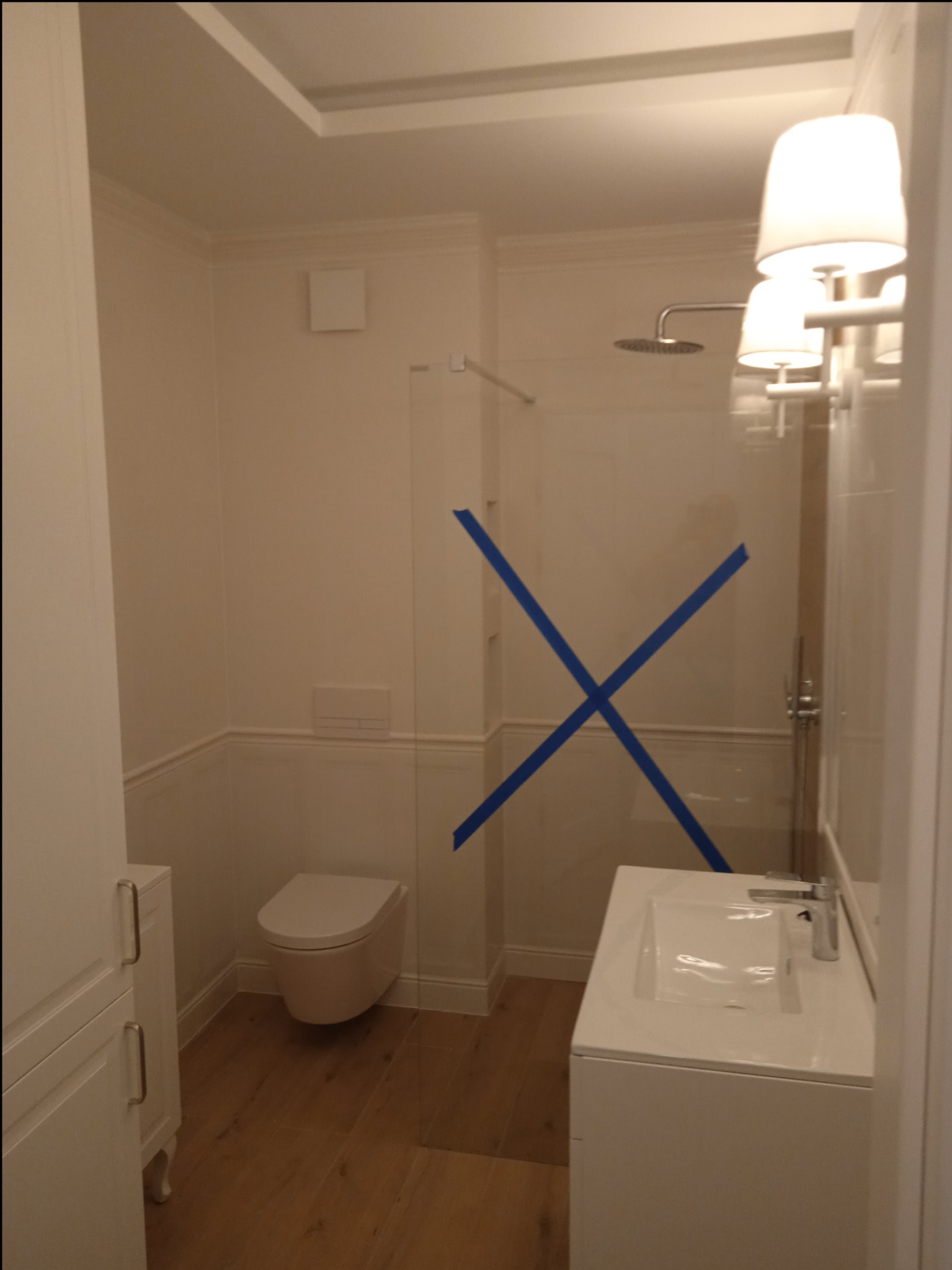 Nowoczesna, biała łazienka z kabiną prysznicową zabezpieczoną niebieską taśmą, wiszącą toaletą, umywalką w zabudowie i drewnopodobną podłogą.