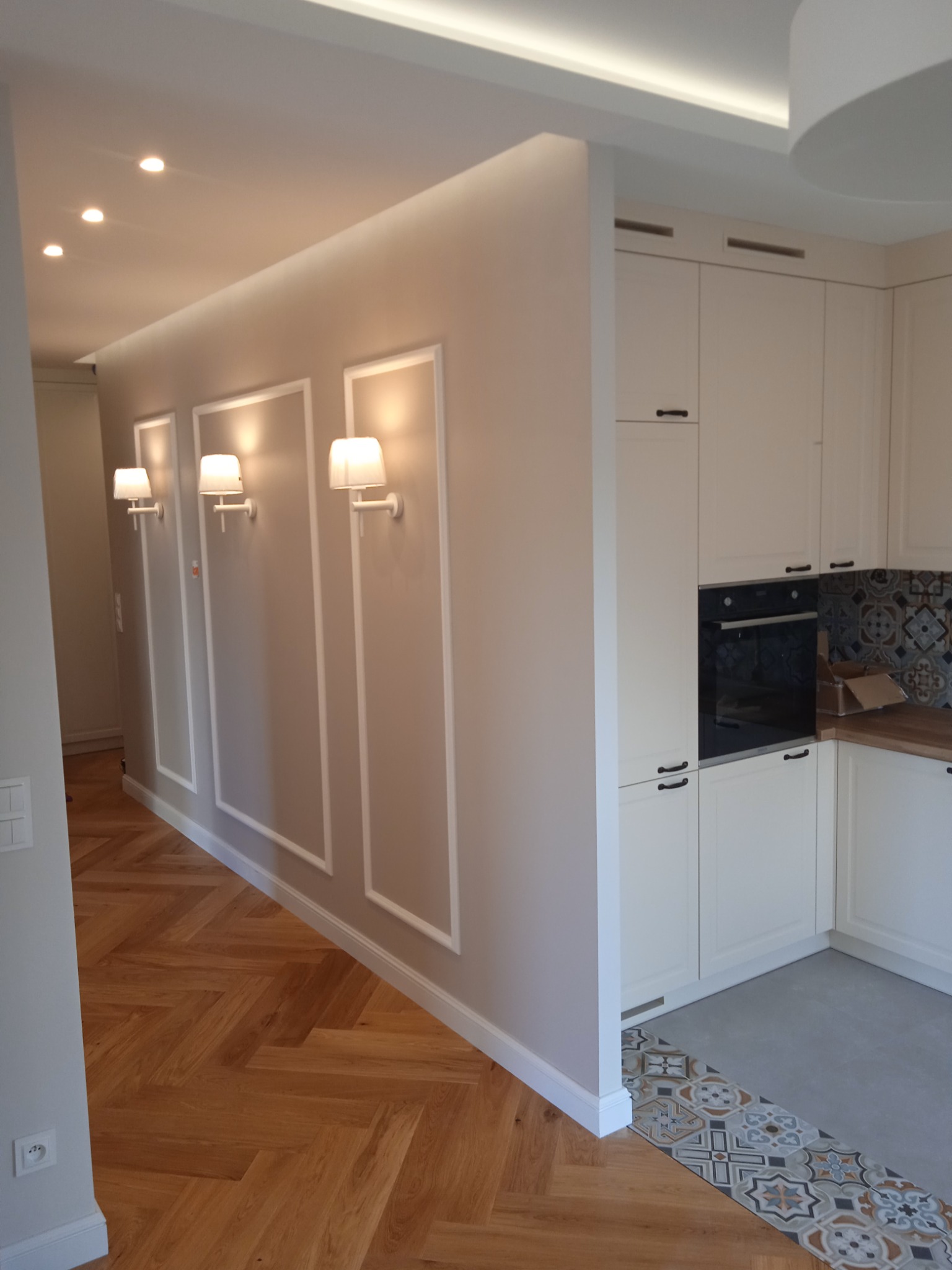 Nowoczesne wnętrze: parkiet w jodełkę, ściana z dekoracyjnymi ramami i kinkietami, oraz fragment kuchni z białymi szafkami i ozdobnymi płytkami.