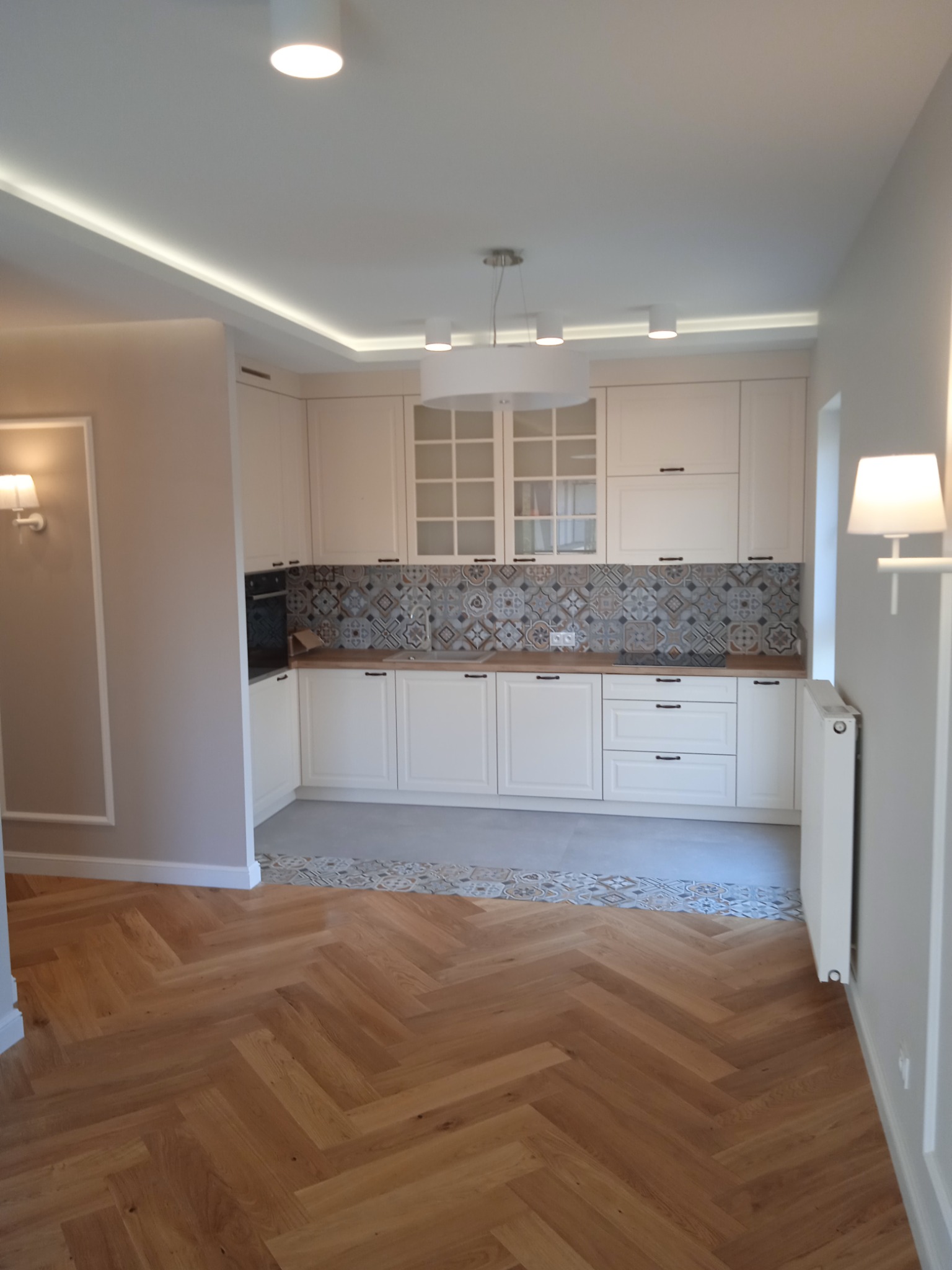 Jasna kuchnia z drewnianą podłogą ułożoną w jodełkę, dekoracyjnymi płytkami nad blatem i oświetleniem LED w suficie podwieszanym.