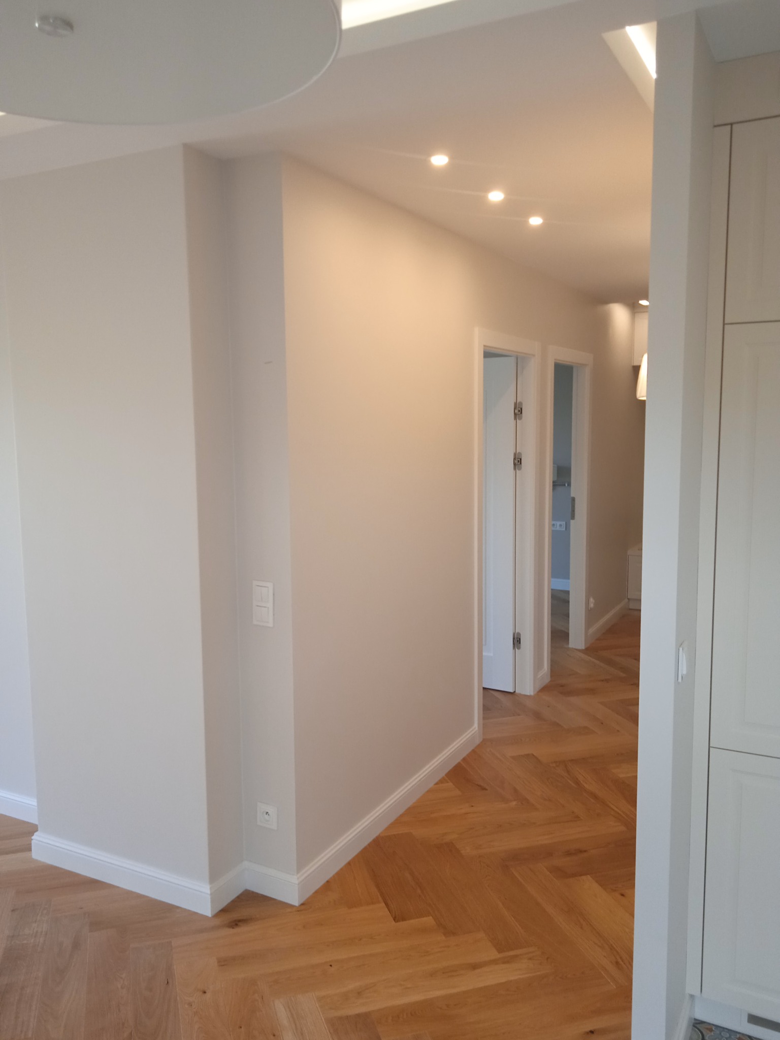 Jasne wnętrze z parkietem ułożonym w jodełkę, świeżo pomalowanymi ścianami w odcieniach bieli i otwartymi drzwiami do kolejnych pomieszczeń.