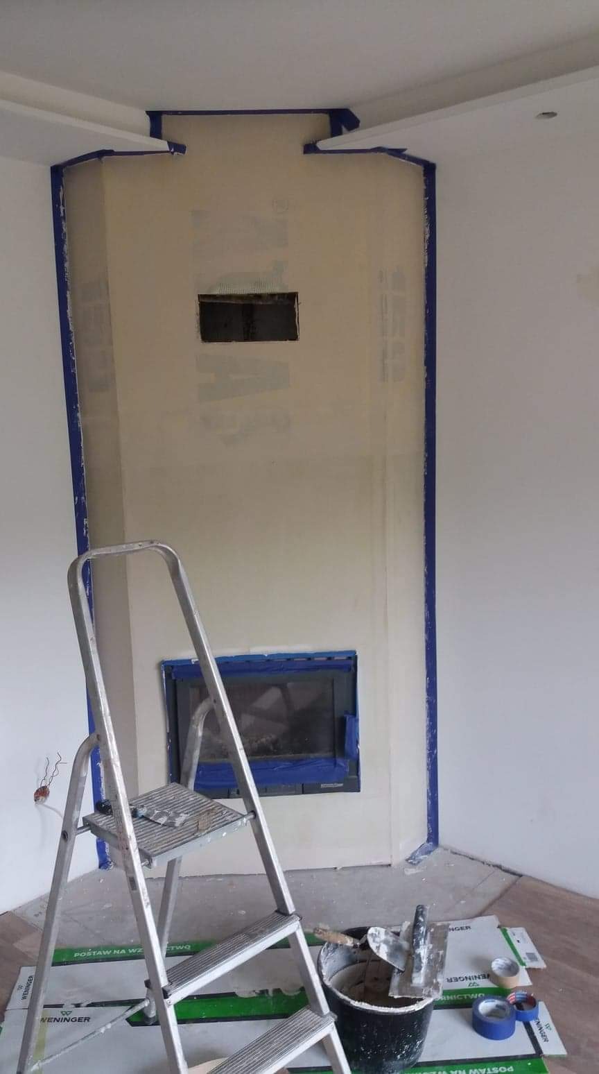 Narożny kominek obudowany płytami, zabezpieczony taśmą malarską, w trakcie szpachlowania, widoczna drabina i narzędzia.
