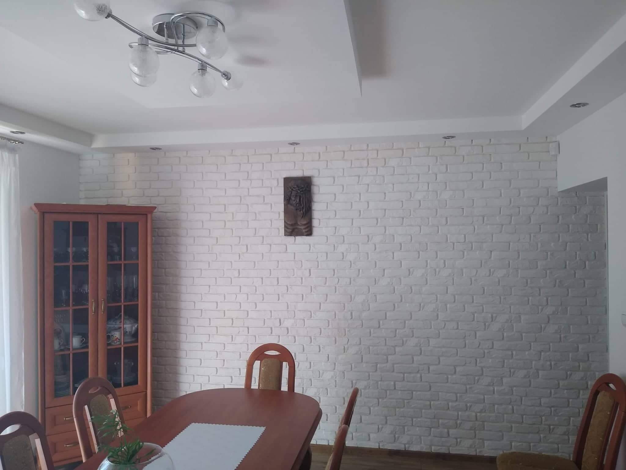 Biała ściana z cegieł dekoracyjnych w jadalni, oświetlona lampą sufitową, z drewnianymi meblami i dekoracją ścienną.