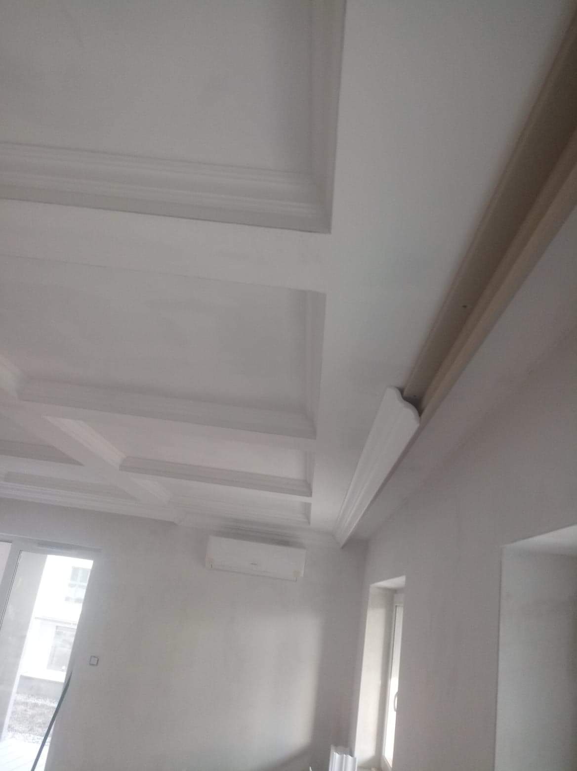 Wykończony sufit z dekoracyjnymi elementami gipsowymi, widoczny klimatyzator na ścianie i okno w tle.