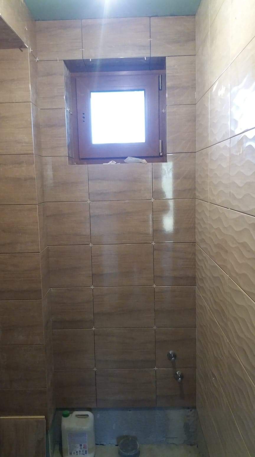 Ściana łazienki w trakcie wykańczania brązowymi płytkami imitującymi drewno, widoczne krzyżyki dystansowe, okno z drewnianą ramą oraz instalacja wodna, na podłodze pojemnik z klejem do płytek.