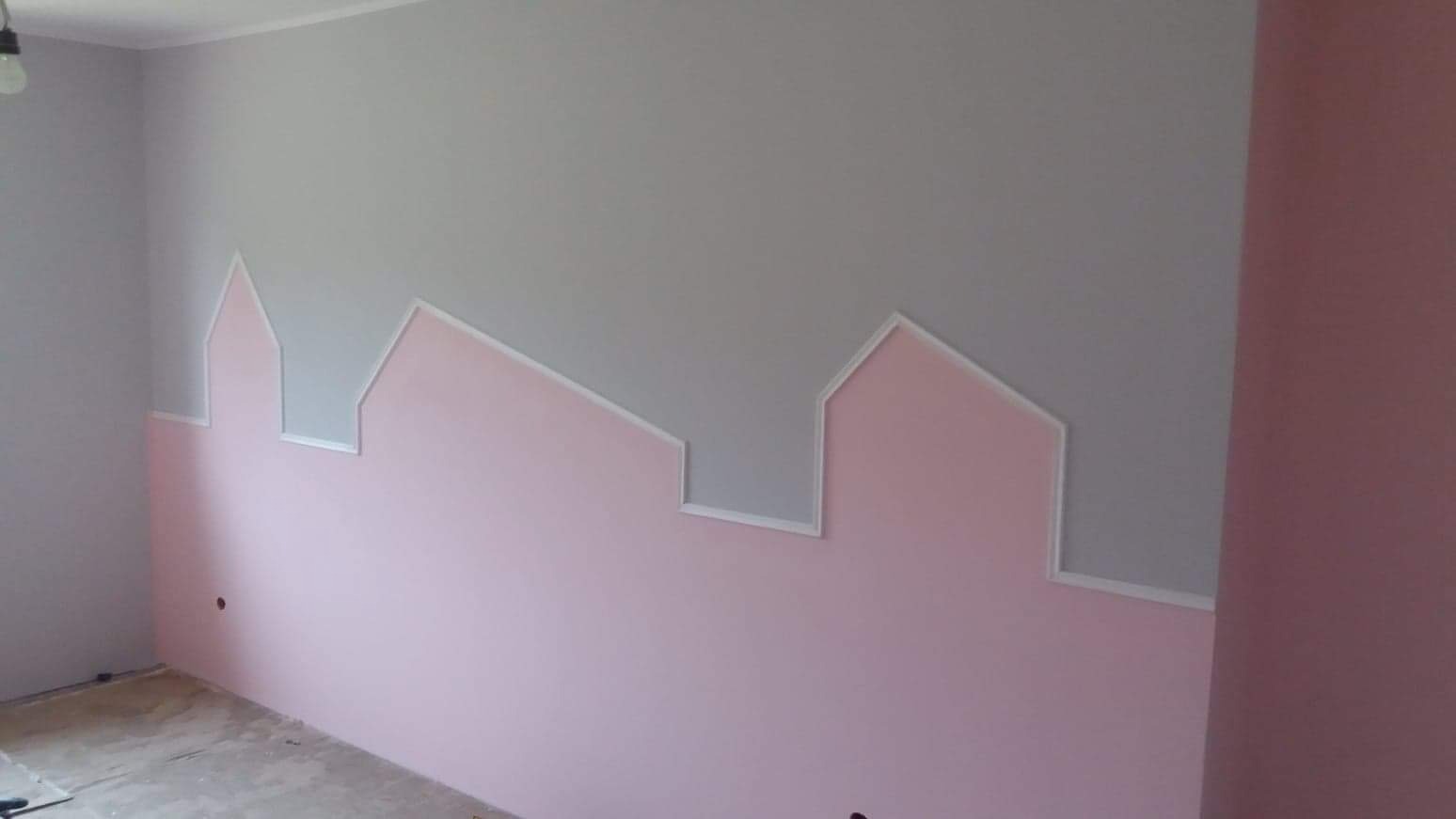 Ściana w pokoju dziecięcym pomalowana na szaro i różowo, z dekoracyjnym obramowaniem w kształcie domków, tworzącym iluzję panoramy miasta.