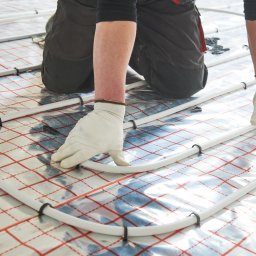 Elektryczna pomarańcza sp. z o.o. - Instalator w białych rękawicach układa rury Purmo ogrzewania podłogowego na folii z czerwoną siatką, widoczne czarne uchwyty mocujące.
