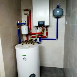 Biały, cylindryczny zasobnik ciepłej wody użytkowej Gree, podłączony rurami z tworzywa w kolorach czerwonym i niebieskim do wiszącego, białego kotła gazowego w kotłowni wyłożonej beżowymi płytkami...