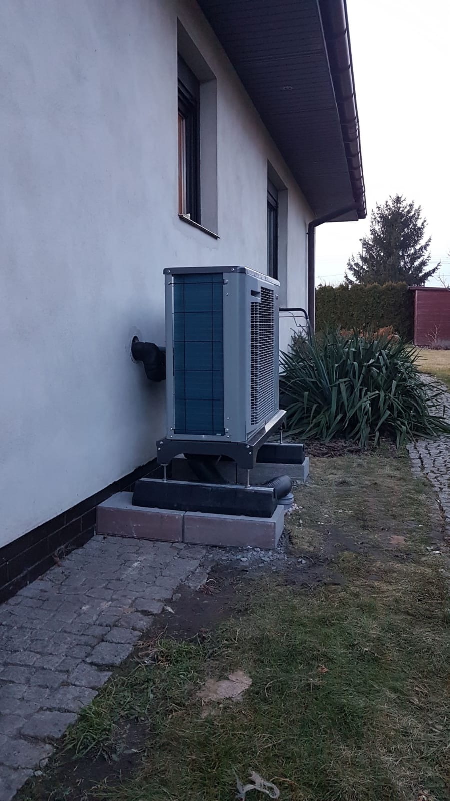 Pompa ciepła Nibe monoblok, zamontowana na zewnątrz budynku na betonowej podstawie, z widocznymi rurami odprowadzającymi wodę, na tle trawnika i krzewów.