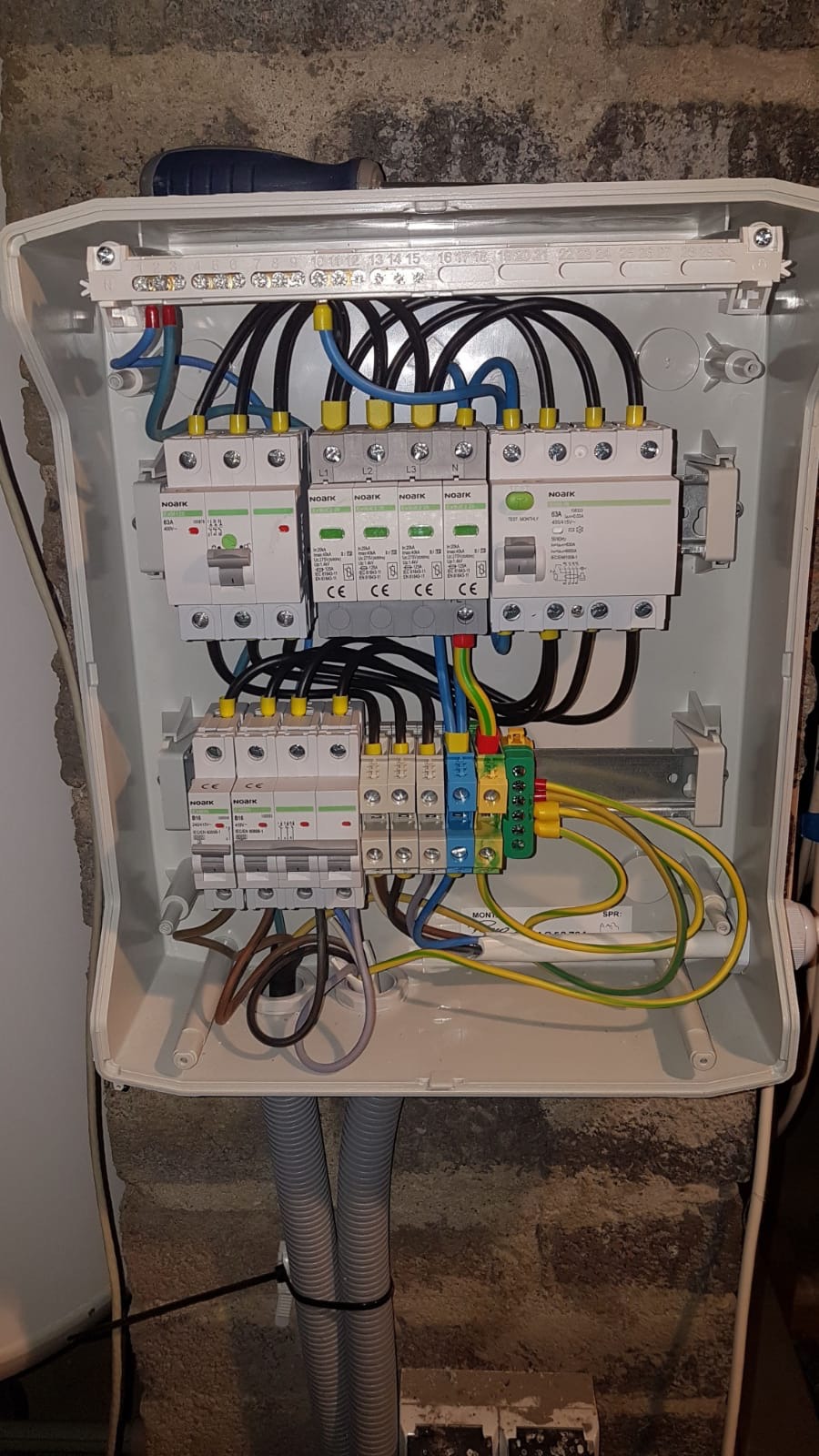 Otwarta skrzynka elektryczna z zainstalowanymi bezpiecznikami i okablowaniem, widoczne kable w kolorach czarnym, niebieskim, żółto-zielonym, oraz szary peszel doprowadzający przewody do skrzynki.