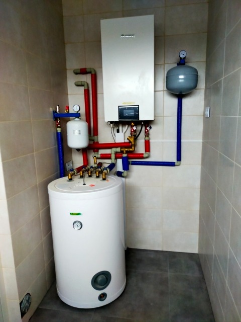 Biały, cylindryczny zasobnik ciepłej wody użytkowej Gree, podłączony rurami z tworzywa w kolorach czerwonym i niebieskim do wiszącego, białego kotła gazowego w kotłowni wyłożonej beżowymi płytkami...