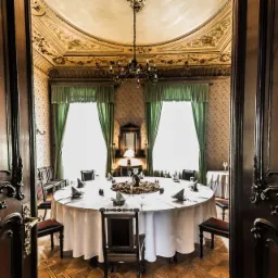 Elegancka sala jadalna z okrągłym stołem nakrytym białym obrusem, widok przez ozdobne, ciemne drzwi, żyrandol i zdobiony sufit.