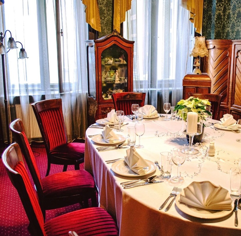 Elegancko nakryty okrągły stół z białym obrusem, zastawą, złożonymi serwetkami i szklankami, otoczony czerwonymi krzesłami w pomieszczeniu z dekoracyjnymi meblami i zasłonami.