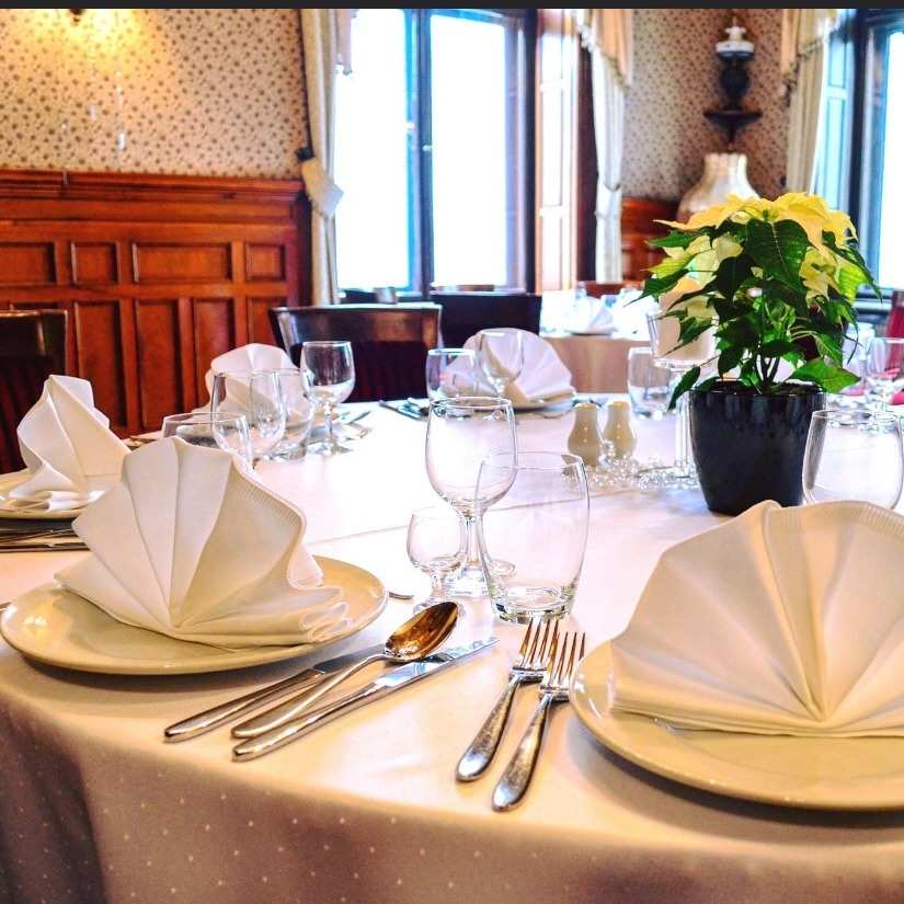 Elegancko nakryty stół z białym obrusem w kropki, talerze z serwetkami złożonymi w wachlarz, sztućce, kieliszki, solniczka i pieprzniczka oraz doniczka z białą poinsecją, w tle boazeria i tapeta...