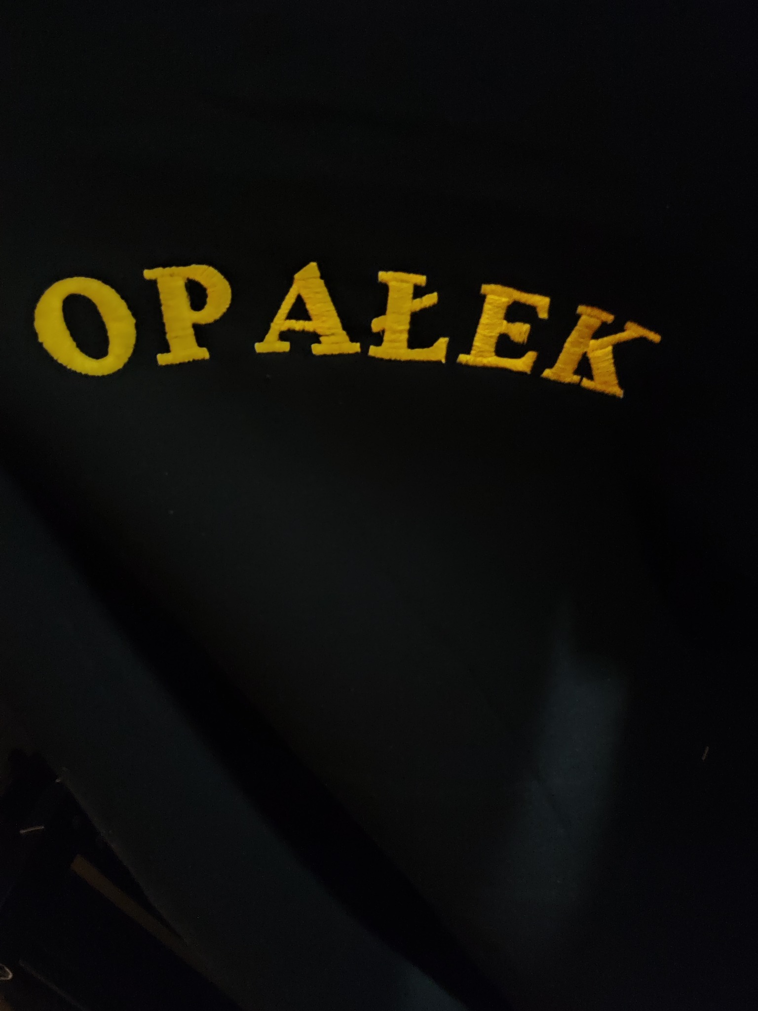 Ciemne tło z wyhaftowanym, żółtym napisem 'OPAŁEK' w górnej części kadru.