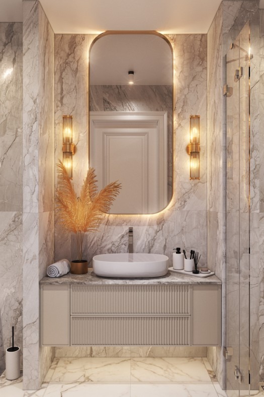 Elegancka łazienka z marmurowymi ścianami, owalną umywalką nablatową, szafką z ryflowanymi frontami, podświetlanym lustrem w złotej ramie i złotymi kinkietami.