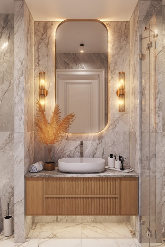 Elegancka łazienka z marmurowymi ścianami, owalną umywalką nablatową, drewnianą szafką z ryflowanymi frontami, podświetlanym lustrem w złotej ramie i dekoracyjnymi lampami ściennymi.