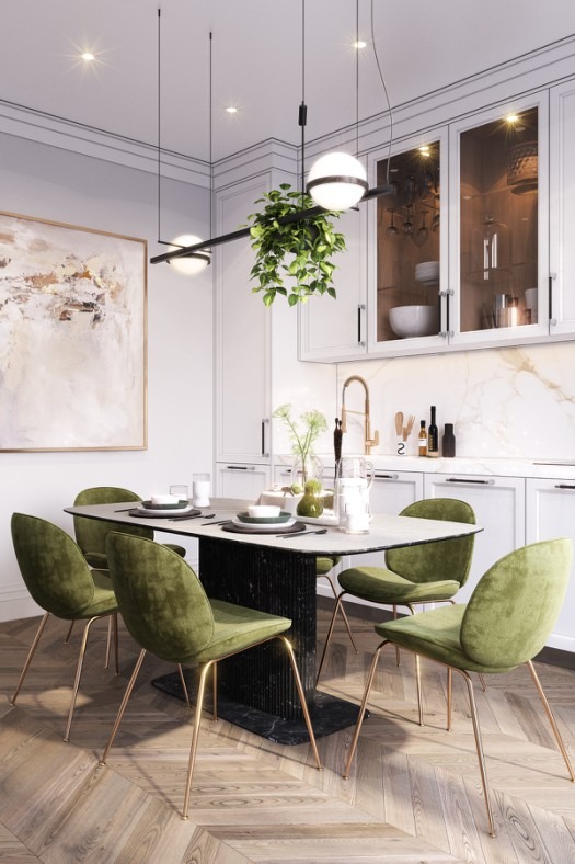 Nowoczesna jadalnia z białymi szafkami, marmurowym blatem, stołem z czarną podstawą i zielonymi krzesłami na złotych nogach, oświetlona wiszącą lampą z rośliną.