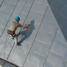 Pracownik w uprzęży, z niebieskim kaskiem, zabezpieczony linami, spryskuje metalowy dach z blachy na rąbek stojący, widok z góry.