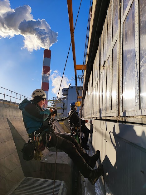 Dwóch alpinistów przemysłowych w kaskach i uprzężach, zwisa na linach z boku budynku przemysłowego z widocznym kominem w tle, wykonując prace na wysokości.