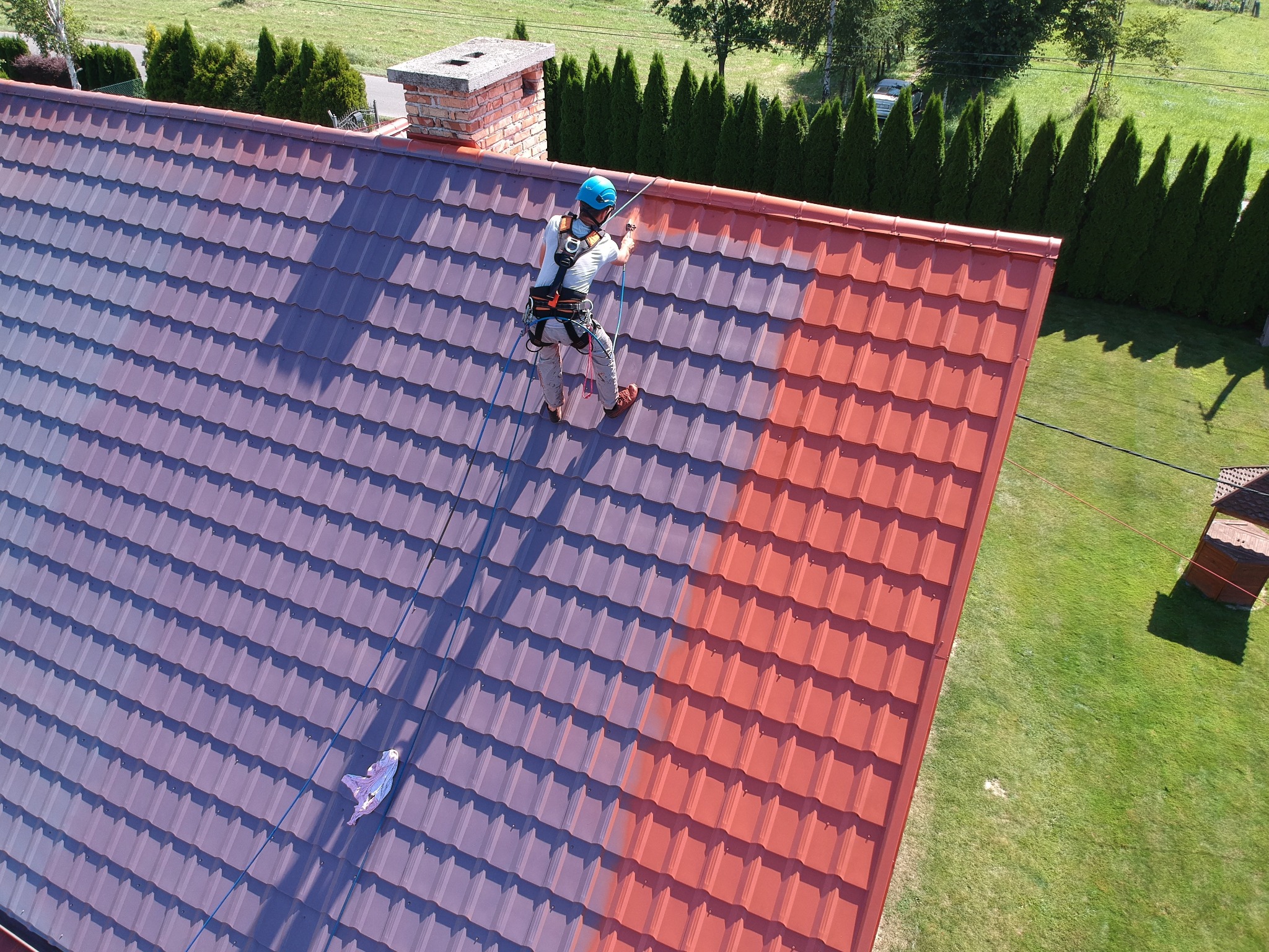 Pracownik w uprzęży, z niebieskim kaskiem, natryskowo nanosi czerwoną farbę na metalową dachówkę, kontrastując z fioletową, niepomalowaną częścią dachu. Widok z góry, na tle zielonego trawnika...