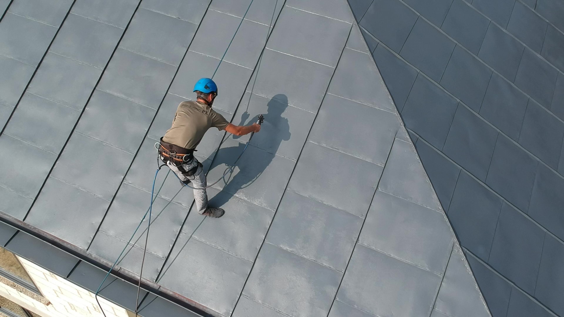 Pracownik w uprzęży, z niebieskim kaskiem, zabezpieczony linami, spryskuje metalowy dach z blachy na rąbek stojący, widok z góry.