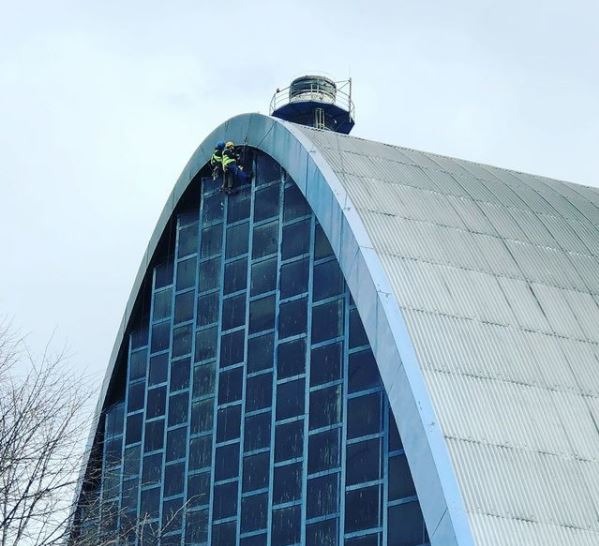 Dwóch pracowników w uprzężach na linach czyści okna na elewacji wysokiego, łukowatego budynku z przeszkloną fasadą i metalowym dachem.