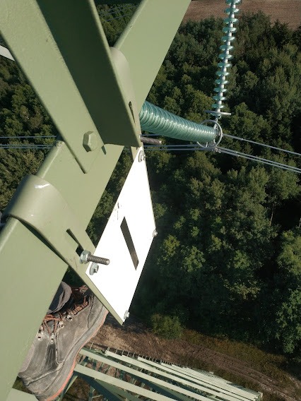 Fragment stalowej konstrukcji wieży energetycznej z widocznym izolatorem łańcuchowym, but pracownika i tabliczka z oznaczeniem, widok z góry na las.