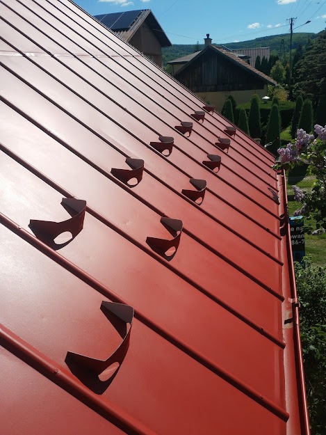Czerwony, blaszany dach z zamontowanymi uchwytami na zabezpieczenia przeciwśniegowe, widok z boku, w tle zabudowa jednorodzinna i zieleń.
