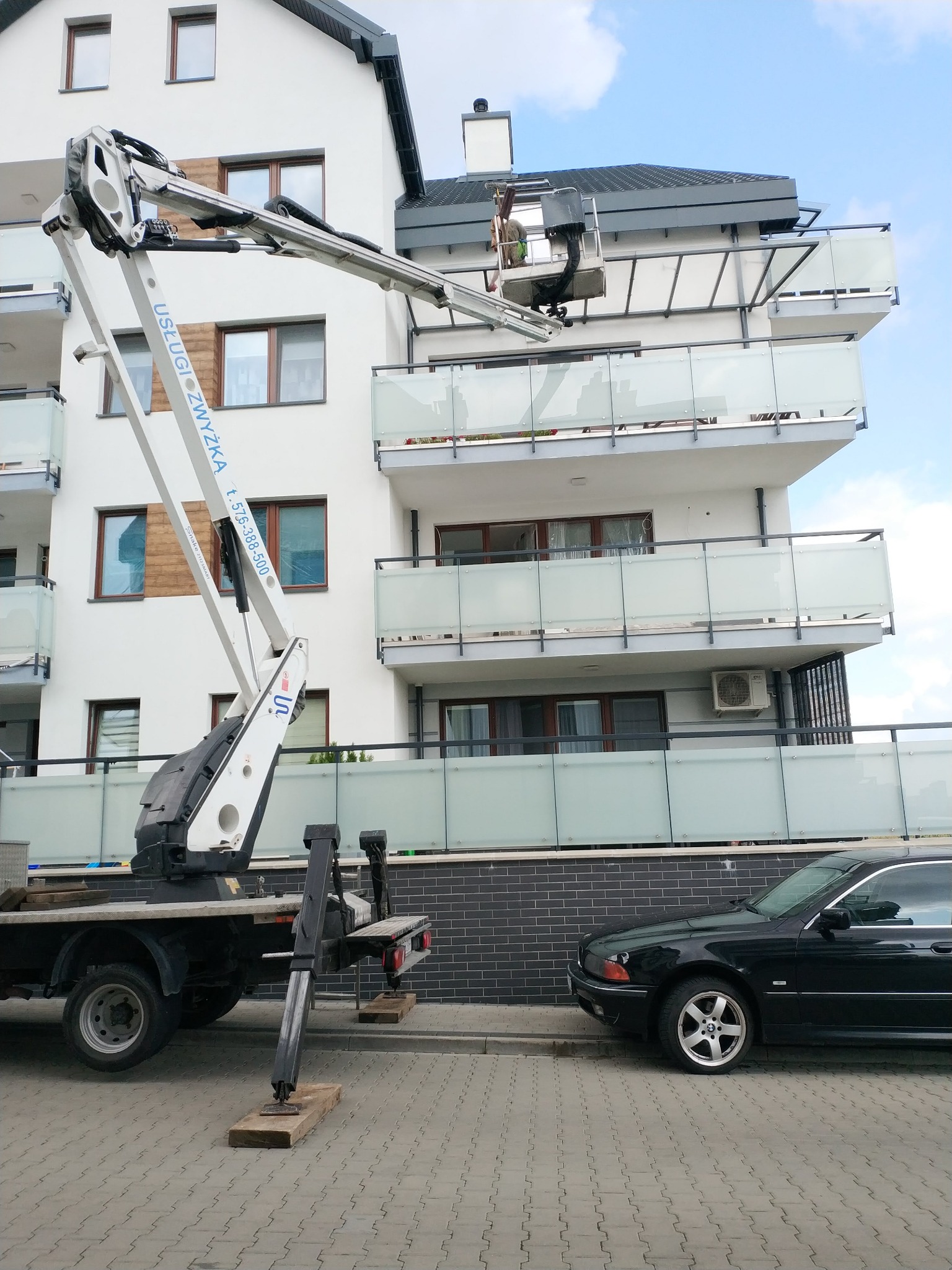 Podnośnik koszowy na tle nowoczesnego budynku z balkonami, pracownik w koszu na wysokości, częściowo widoczny zaparkowany czarny samochód osobowy.