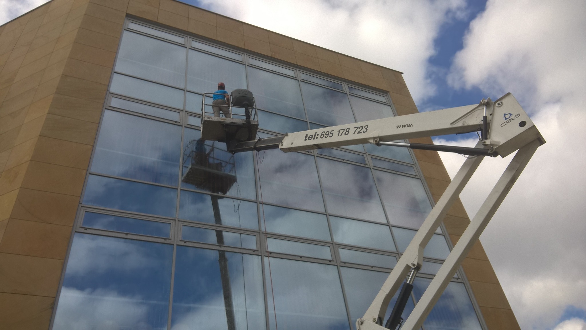 Pracownik w podnośniku koszowym czyści duże okna biurowca, odbijające błękitne niebo z chmurami. Widoczny numer telefonu na ramieniu podnośnika.