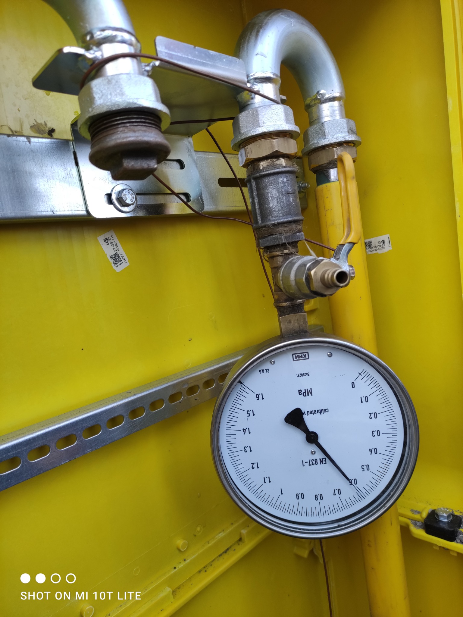 Manometr wskazujący ciśnienie w instalacji gazowej, zamontowany w żółtej skrzynce, widoczne złączki i rury instalacji.