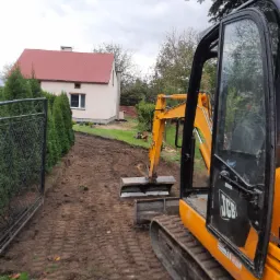 Żółta minikoparka JCB z otwartą kabiną na działce z wykopanym pasem ziemi, widoczny dom z czerwoną dachówką i rząd tuj przy ogrodzeniu.