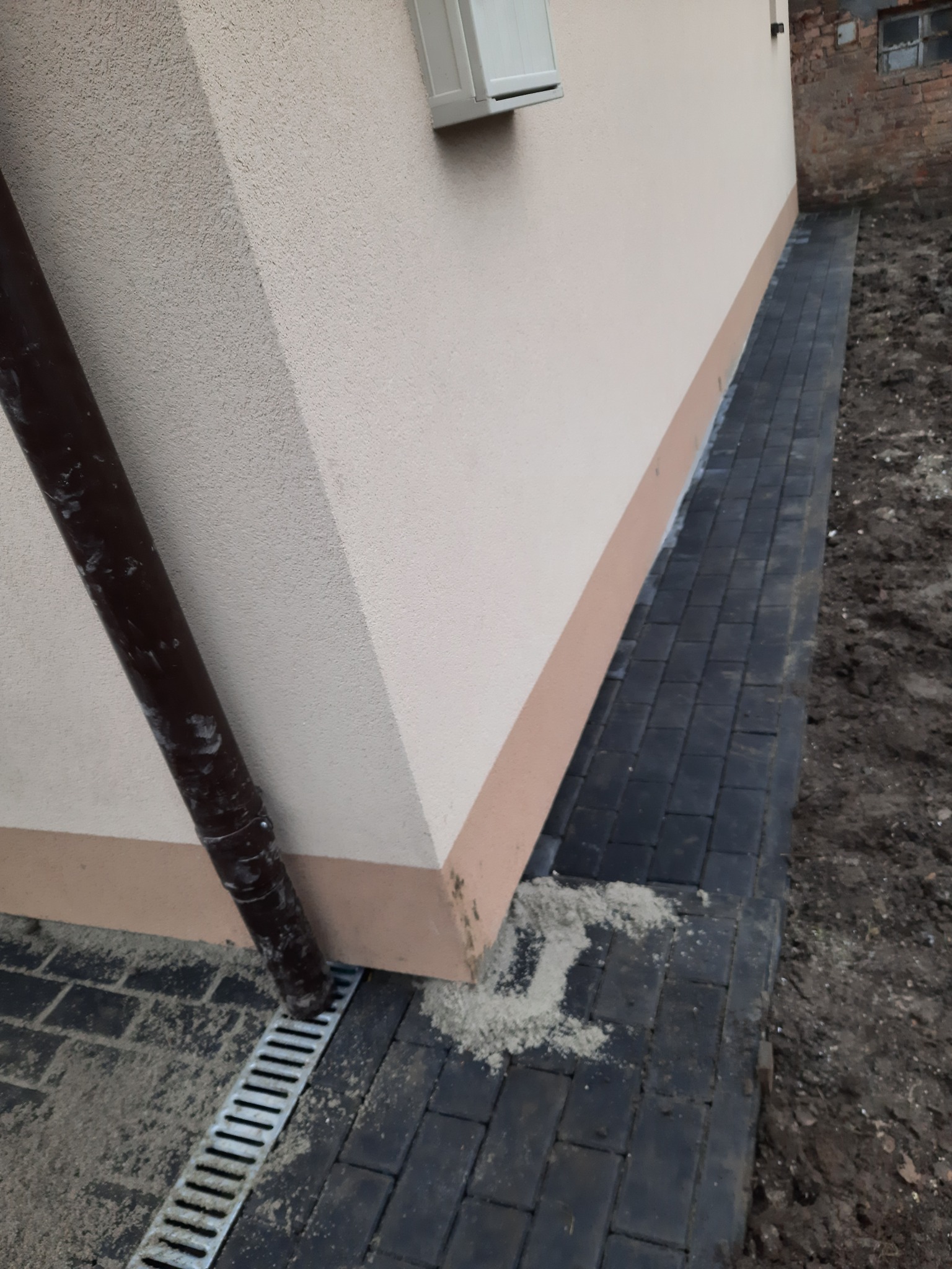 Ciemna kostka brukowa ułożona wzdłuż ściany budynku z zamontowanym odpływem liniowym i rynną spustową, widoczne resztki piasku na kostce.