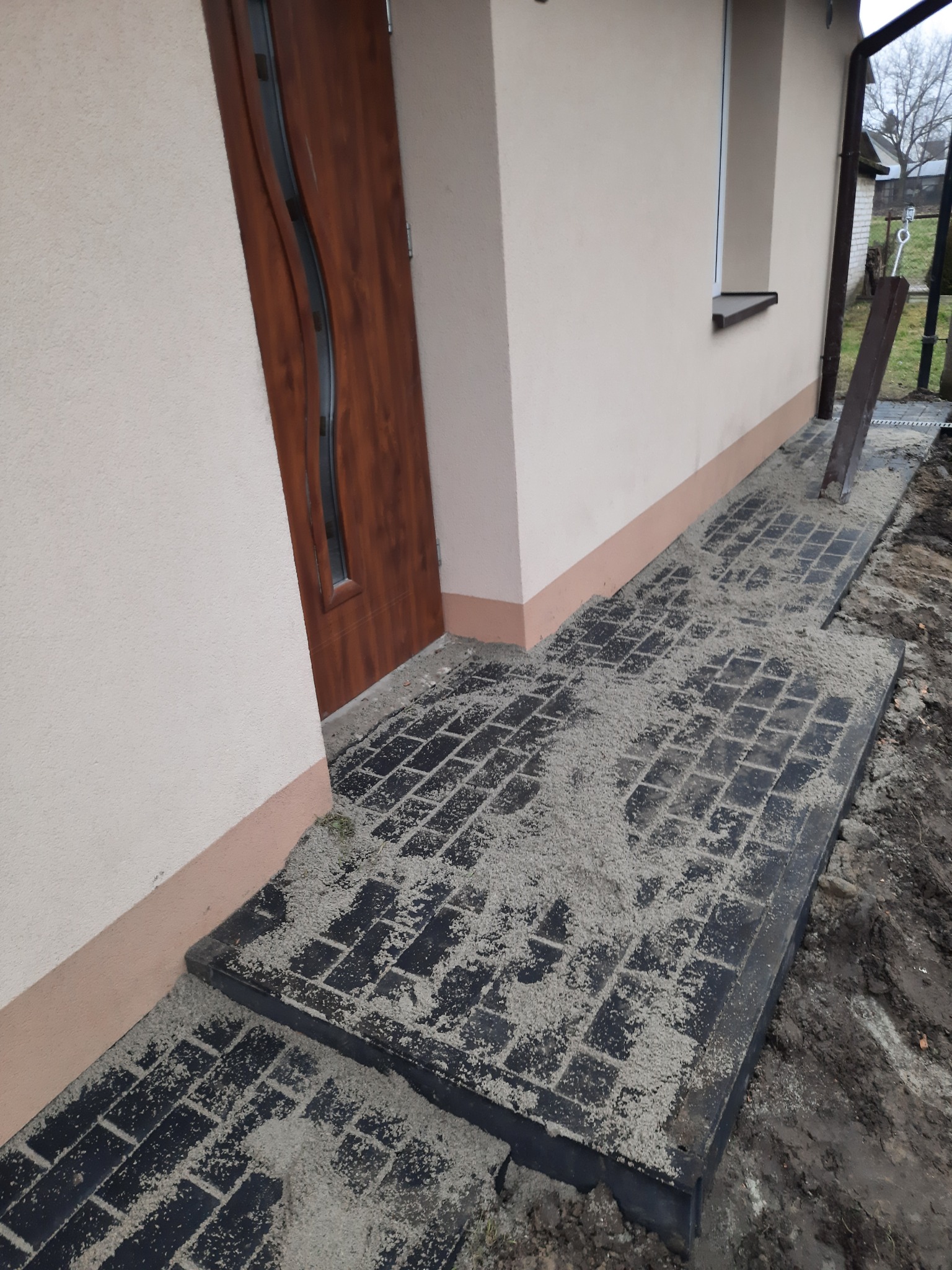 Ciemna kostka brukowa ułożona jako chodnik przed budynkiem, zasypana piaskiem kwarcowym, widoczne obrzeża chodnika.