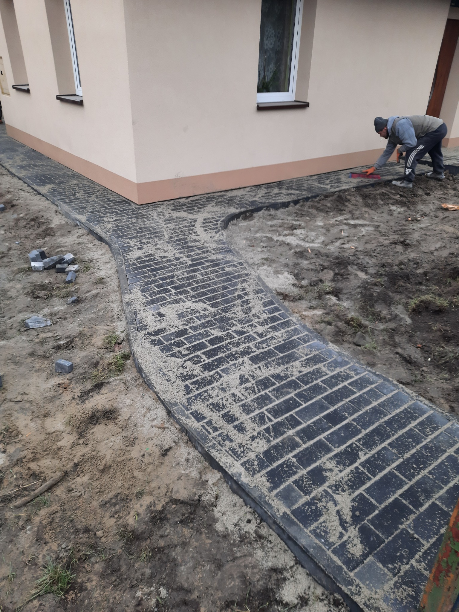 Pracownik układa ciemnoszarą kostkę brukową wokół jasnobeżowego domu z białymi oknami, używając czerwonej gumowej packi do wyrównywania powierzchni.