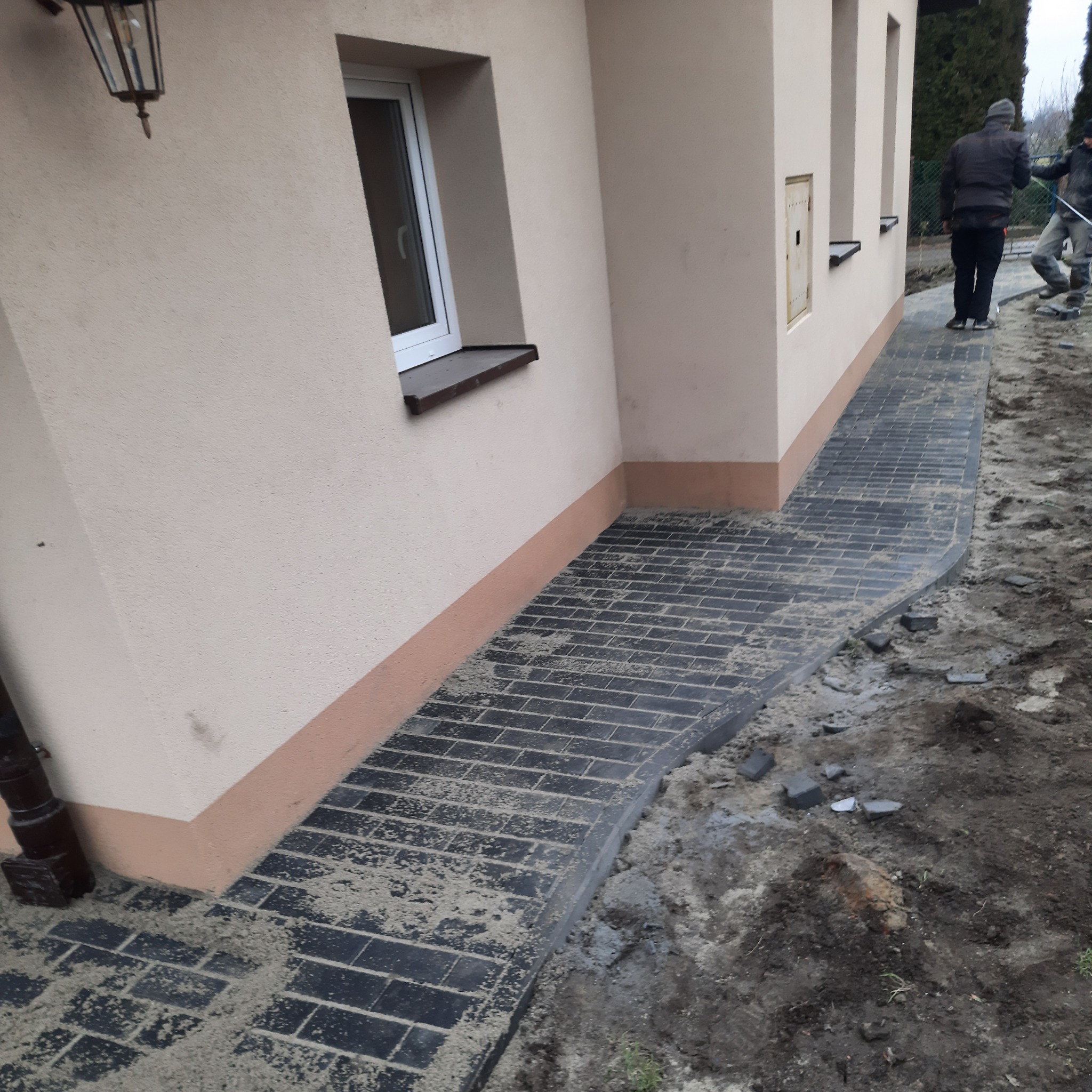 Ułożona ścieżka z ciemnej kostki brukowej przy budynku, widoczni dwaj pracownicy w tle, teren wokół jeszcze niezagospodarowany.