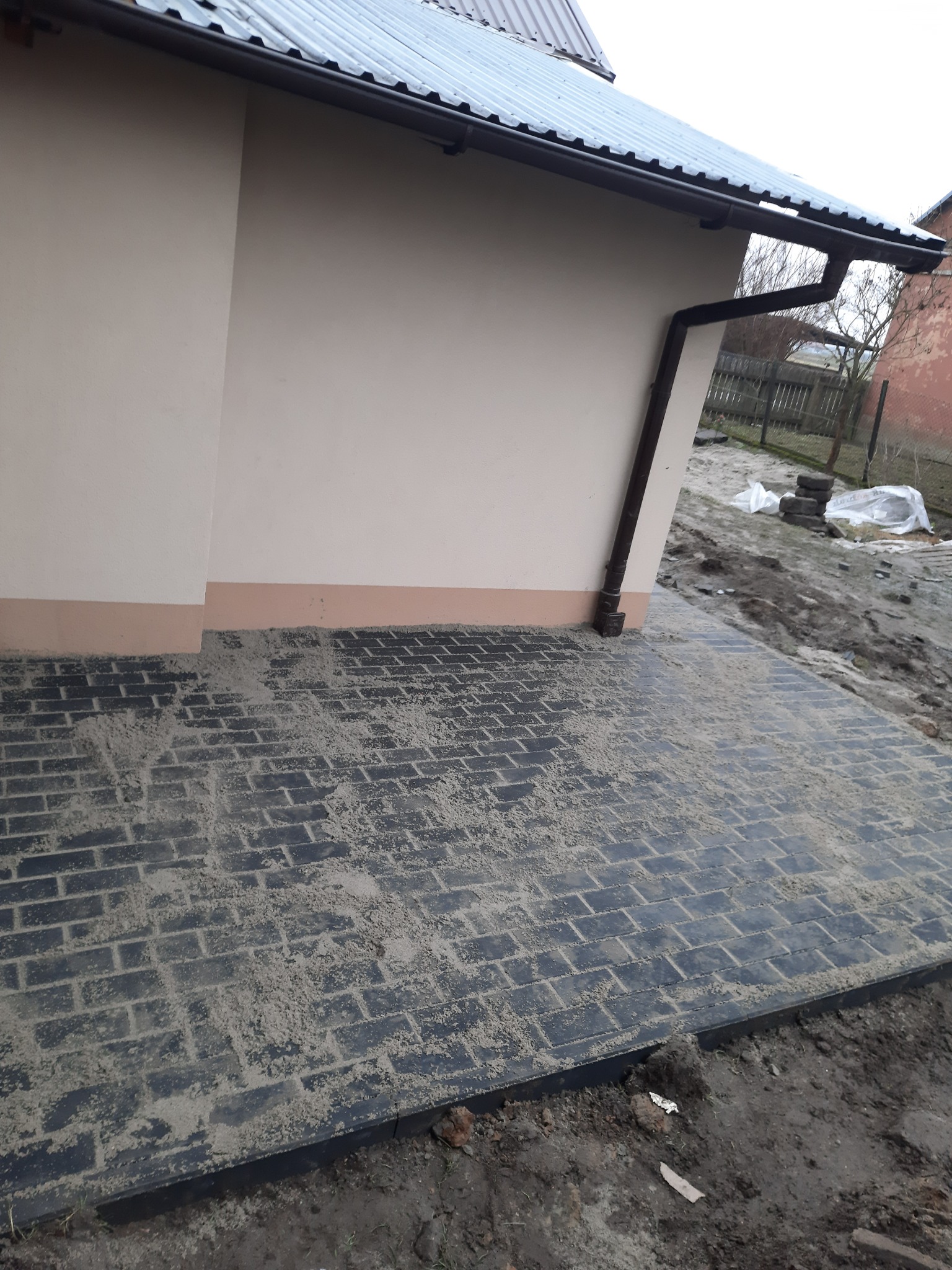 Nowo ułożona, ciemna kostka brukowa przy ścianie budynku z rynną, zasypana piaskiem do wypełnienia szczelin, widoczne nierówności terenu w tle.
