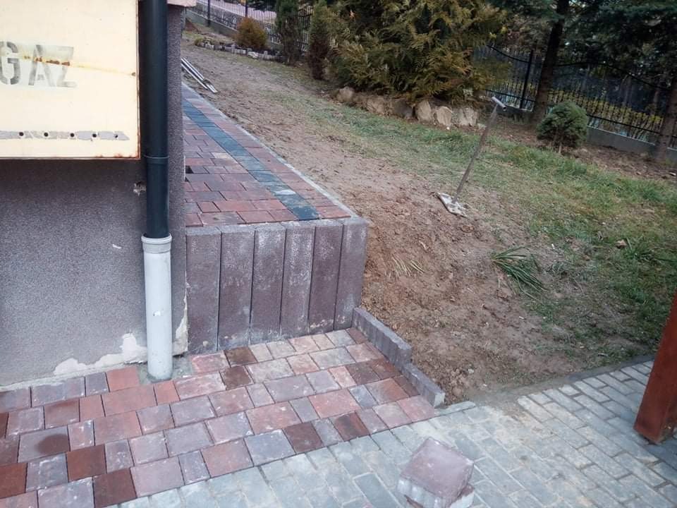 Ułożona kostka brukowa w odcieniach czerwieni i szarości, tworząca ścieżkę i schody przed budynkiem, z widocznym fragmentem elewacji i rurą spustową, na tle ziemi i zieleni.