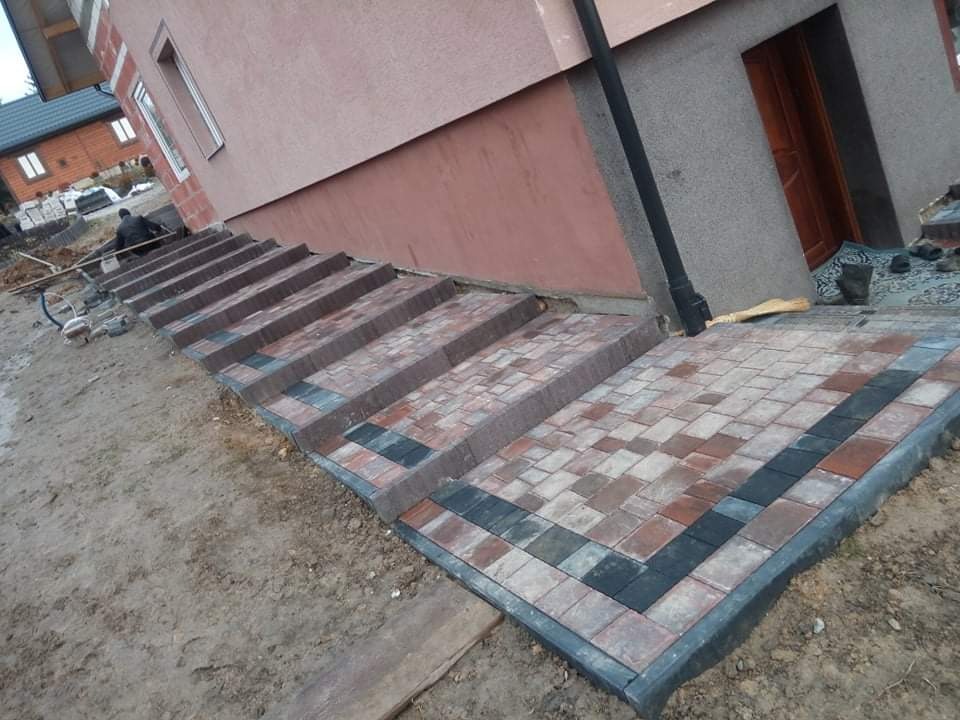 Ułożona kostka brukowa w kolorach brązu, szarości i czerni tworząca schody i chodnik przed budynkiem, widoczny fragment elewacji budynku i pracownik w oddali.